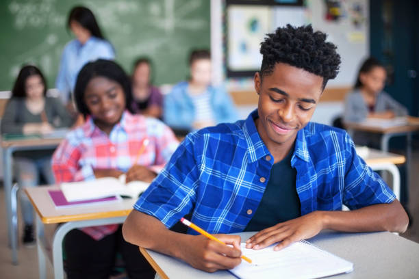 Smiling male student writing in classroom. Teenage boy is concentrating at high school. He is sitting at desk.